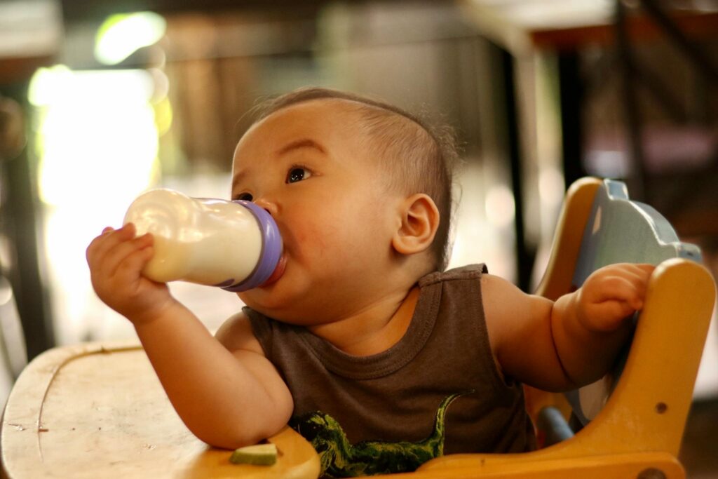 Cute baby in a high chair drinking milk from a bottle indoors, showcasing innocence and nourishment.
