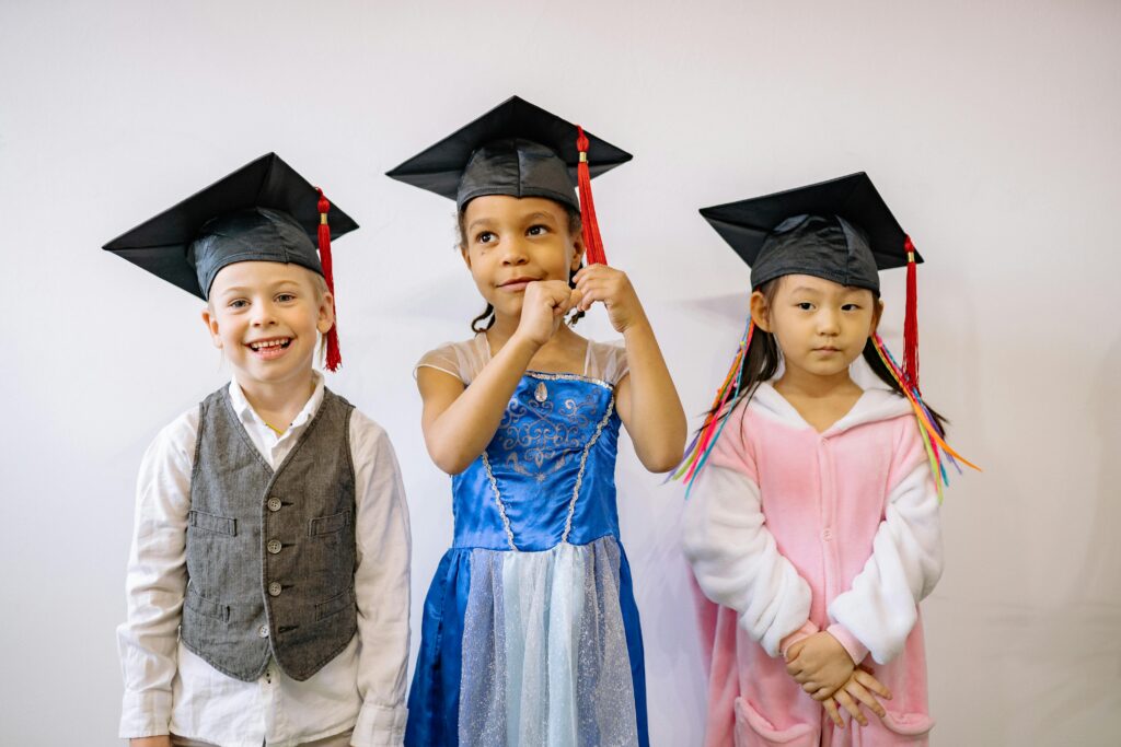 Three children in graduation caps express pride and joy at their preschool graduation ceremony.